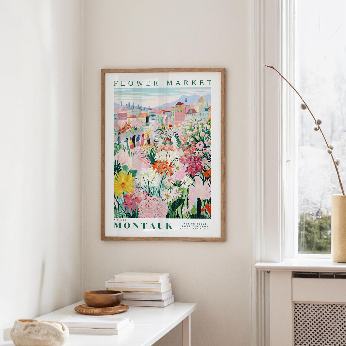 a framed poster of a colorful floral scene hanging on a wall, with a white table and a vase of flowers in the foreground.