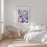 a bedroom with a large poster of the Brisbane Flower Market hanging on the wall. The poster has a colorful illustration of the city skyline and various flowers. The room has a white bed, a wicker chair, and a small side table. The overall color scheme of the room is white and beige, creating a calm and serene atmosphere.