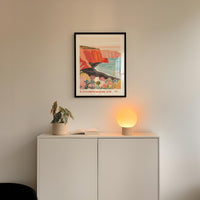 a white cabinet with a potted plant and a lamp on top, set against a white wall with a framed poster of a colorful landscape hanging above it.