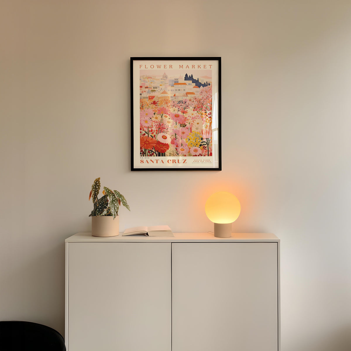 a white cabinet with a potted plant and a lamp on top, set against a wall with a framed poster of a flower market above it.