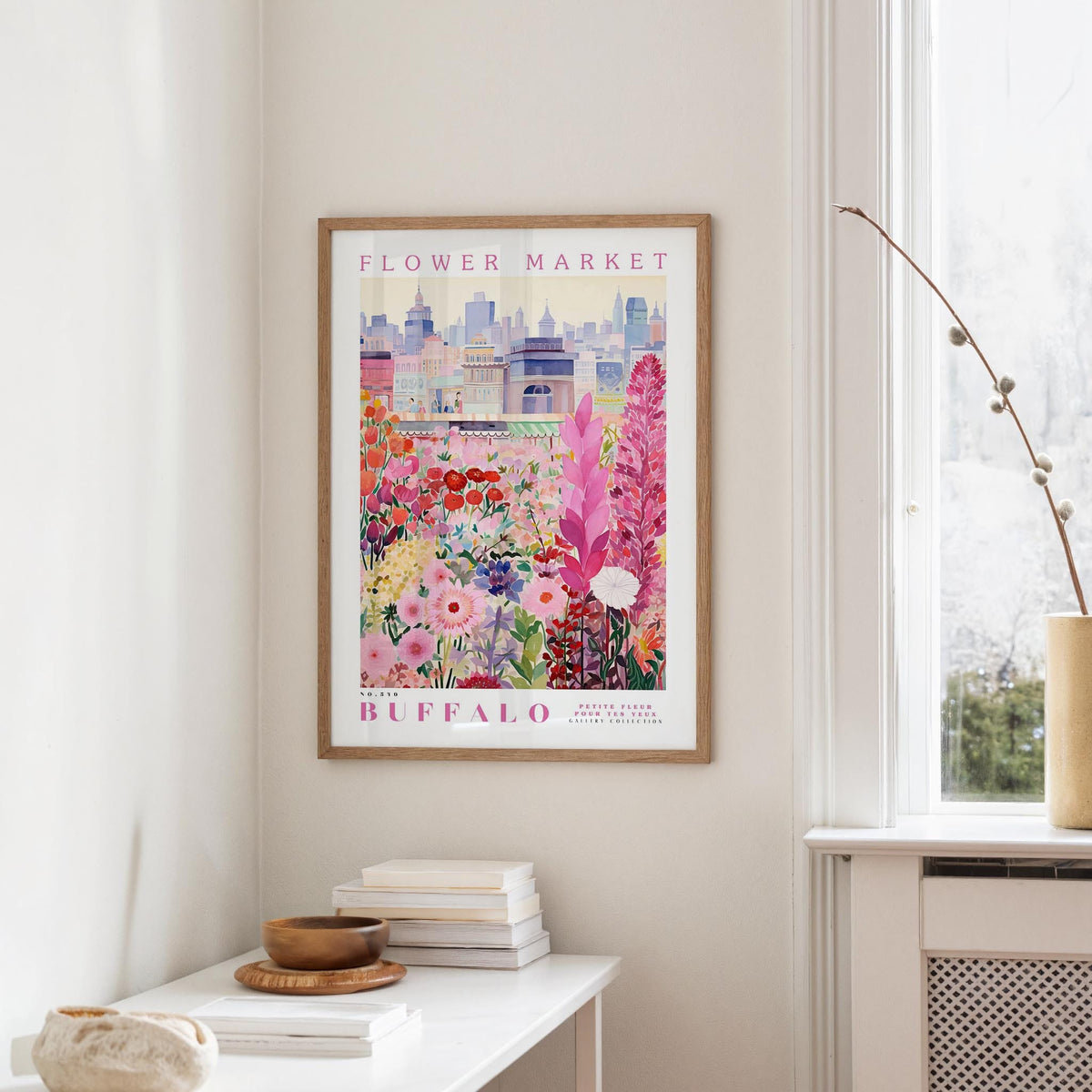 a framed poster of a colorful floral scene hanging on a wall, with a window and a small table with books and a vase in the foreground.