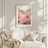 a framed poster of a colorful floral scene hanging on a wall above a white couch with a blanket draped over it.