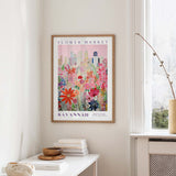 a framed poster of a colorful floral scene hanging on a wall, with a window and a small table with books and a vase in the foreground.