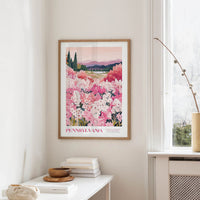 a framed poster of a field of pink flowers hanging on a wall, with a window and a small table with books and a vase nearby.