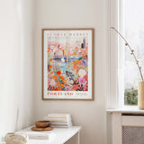 a framed poster of a colorful cityscape with flowers hanging on a wall in a room with a white table and a window.