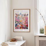 a framed poster of a colorful floral scene hanging on a wall, with a white table and a vase of flowers nearby.