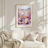 a framed poster of a colorful floral scene hanging on a wall above a white couch with a blanket draped over it.