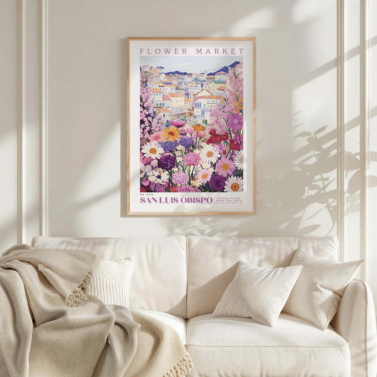 a framed poster of a colorful floral scene hanging on a wall above a white couch with a blanket draped over it.