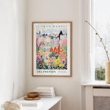 a framed poster of a colorful floral scene hanging on a wall, with a white table and a vase of flowers in the foreground.