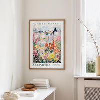 a framed poster of a colorful floral scene hanging on a wall, with a white table and a vase of flowers in the foreground.