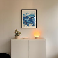 a framed picture of a mountain landscape with a blue and white color scheme, hanging on a wall above a white cabinet. Below the cabinet, there is a small plant and a lamp with a glowing light, creating a cozy and inviting atmosphere.