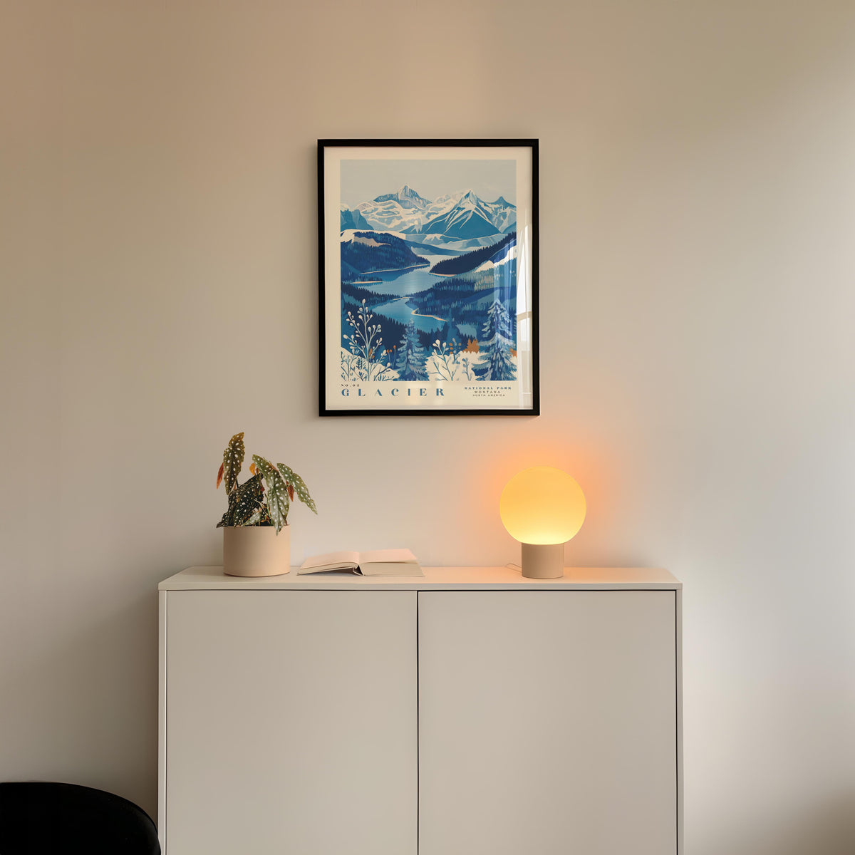 a framed picture of a mountain landscape with a blue and white color scheme, hanging on a wall above a white cabinet. Below the cabinet, there is a small plant and a lamp with a glowing light, creating a cozy and inviting atmosphere.