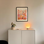 a white cabinet with a potted plant and a lamp on top, set against a wall with a framed poster of a flower market.