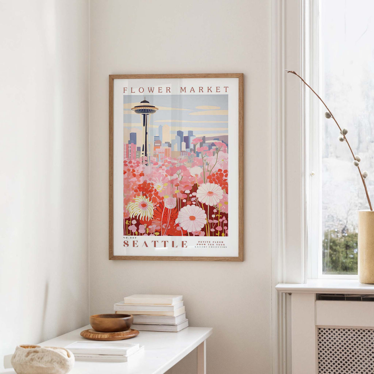 a framed poster of a cityscape with a flower market in the background, hanging on a wall in a room with a window and a small table with books and a vase.