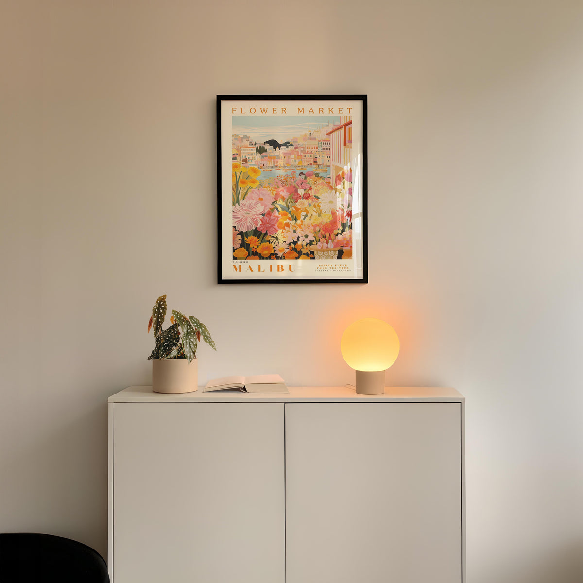 a white cabinet with a potted plant and a lamp on top, set against a wall with a framed poster of a tropical garden hanging above it.