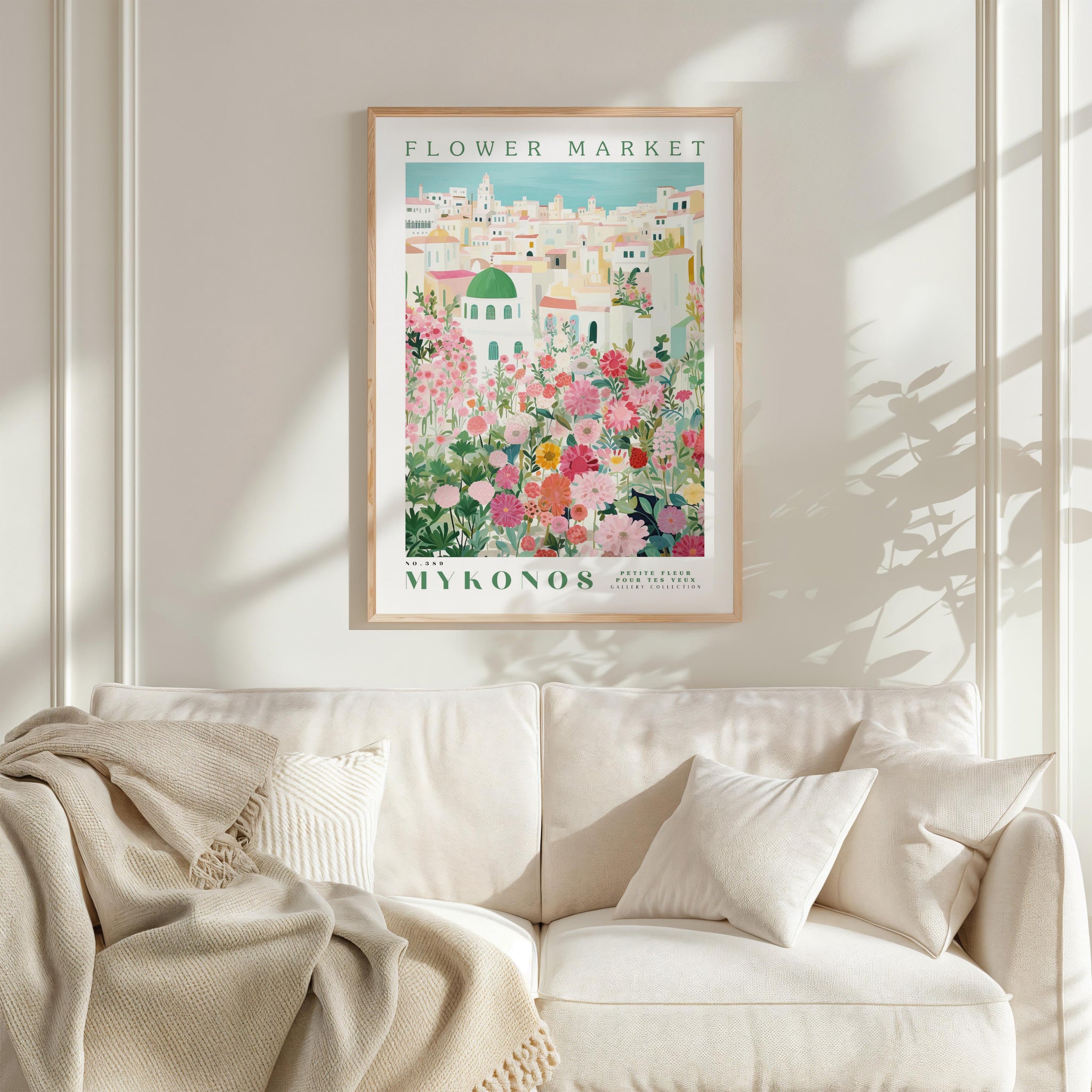 a framed poster of a flower market in Mykonos, Greece, hanging on a white wall above a white couch with a blanket draped over it.