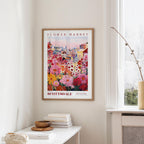 a framed poster of a colorful floral scene hanging on a wall, with a white table and a vase of flowers in the foreground.
