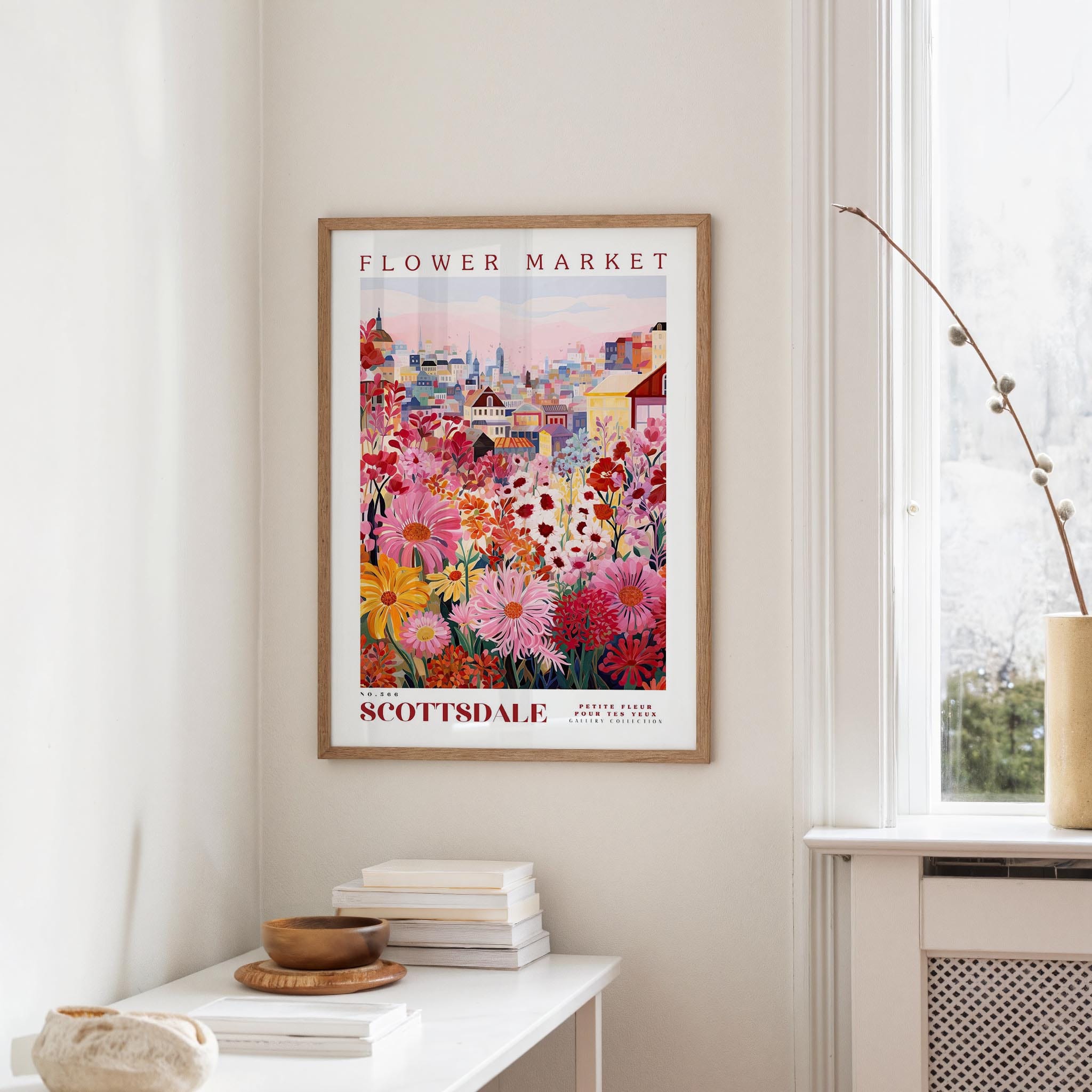 a framed poster of a colorful floral scene hanging on a wall, with a white table and a vase of flowers in the foreground.