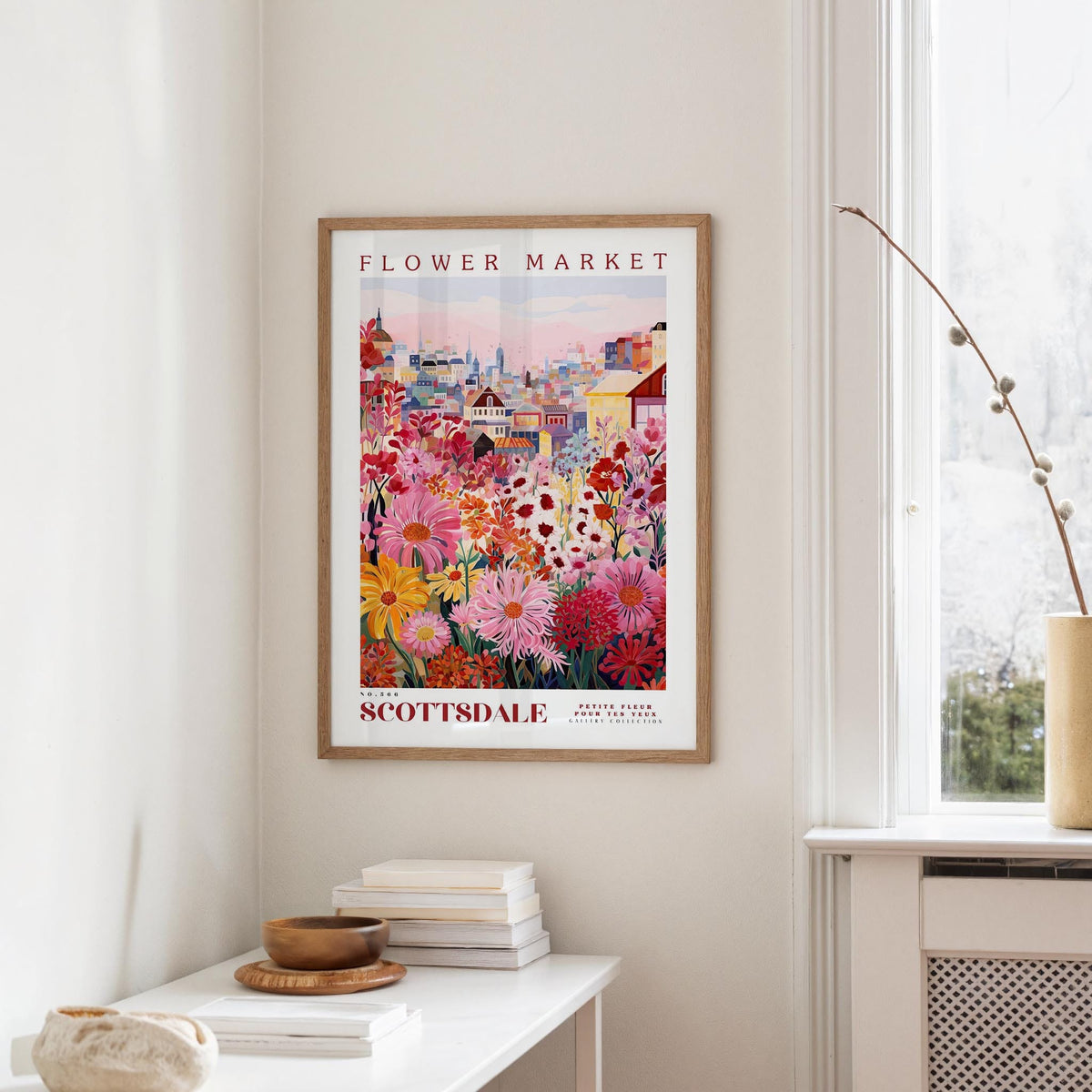 a framed poster of a colorful floral scene hanging on a wall, with a white table and a vase of flowers in the foreground.