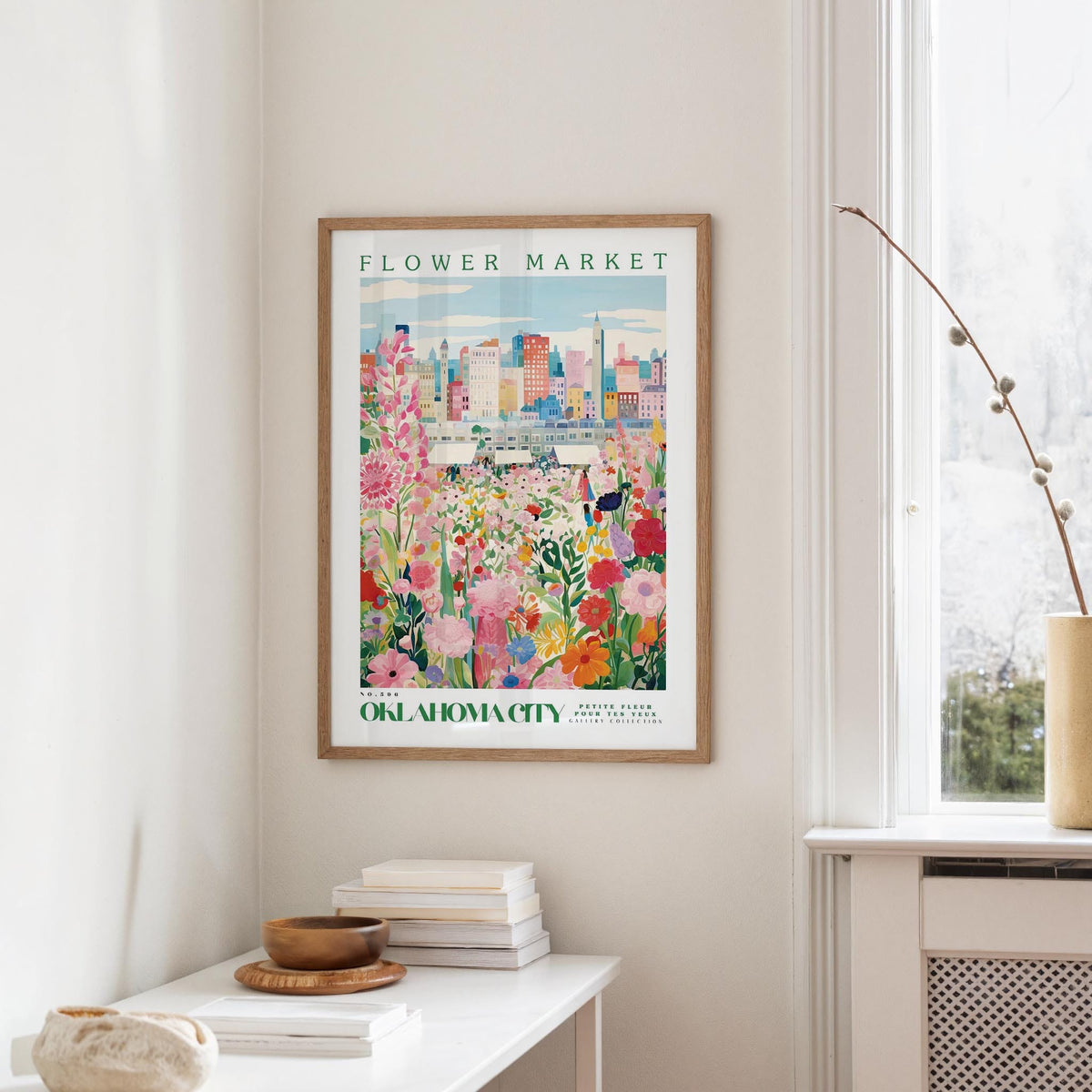 a framed poster of a colorful floral scene hanging on a wall, with a window and a small table with books and a vase in the foreground.