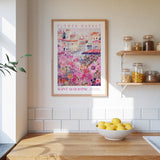 a kitchen scene with a framed poster of a colorful flower market in Saint Augustine, Florida, hanging on the wall above a wooden counter. The poster is surrounded by various kitchen items, such as jars, a bowl of lemons, and a plant.