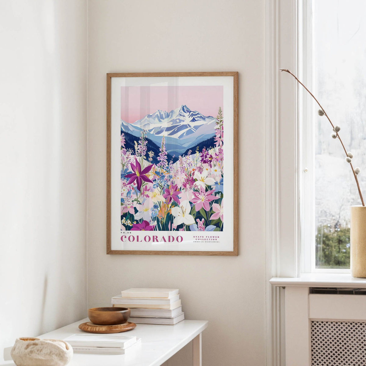 a framed poster of a mountain landscape with a variety of colorful flowers, hanging on a wall in a room with a white table and a window.