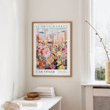 a framed poster of a colorful cityscape with flowers hanging on a wall, next to a window and a small table with books and a bowl.