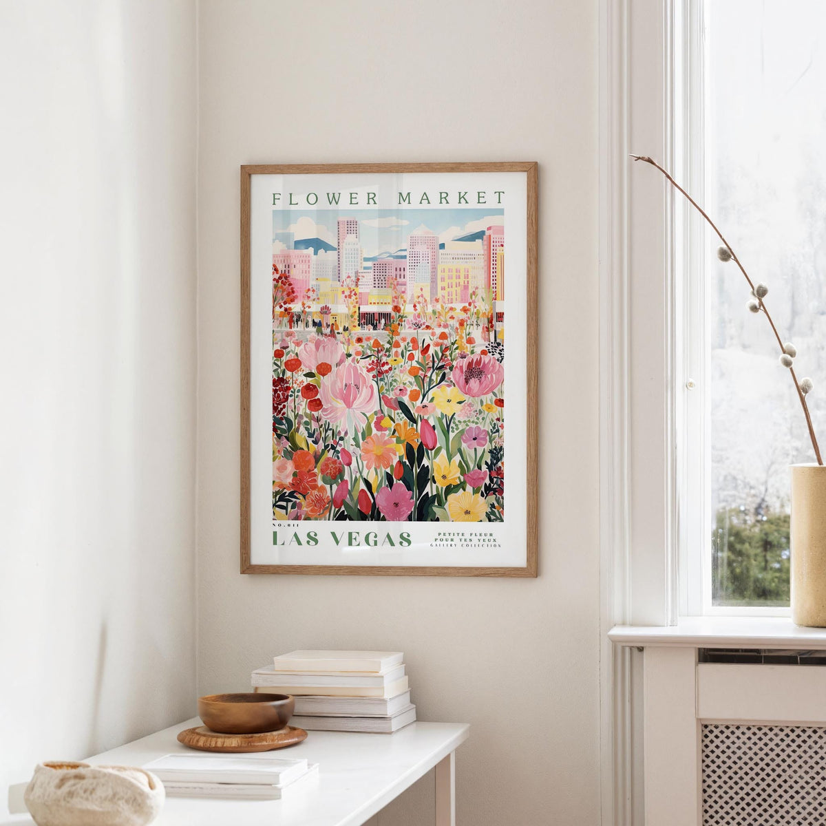 a framed poster of a colorful cityscape with flowers hanging on a wall, next to a window and a small table with books and a bowl.