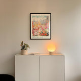 a white cabinet with a potted plant and a lamp on top, set against a wall with a framed poster of a cityscape above it.