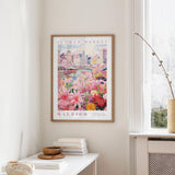 a framed poster of a colorful floral scene hanging on a wall, with a window and a small table with books and a vase in the foreground.