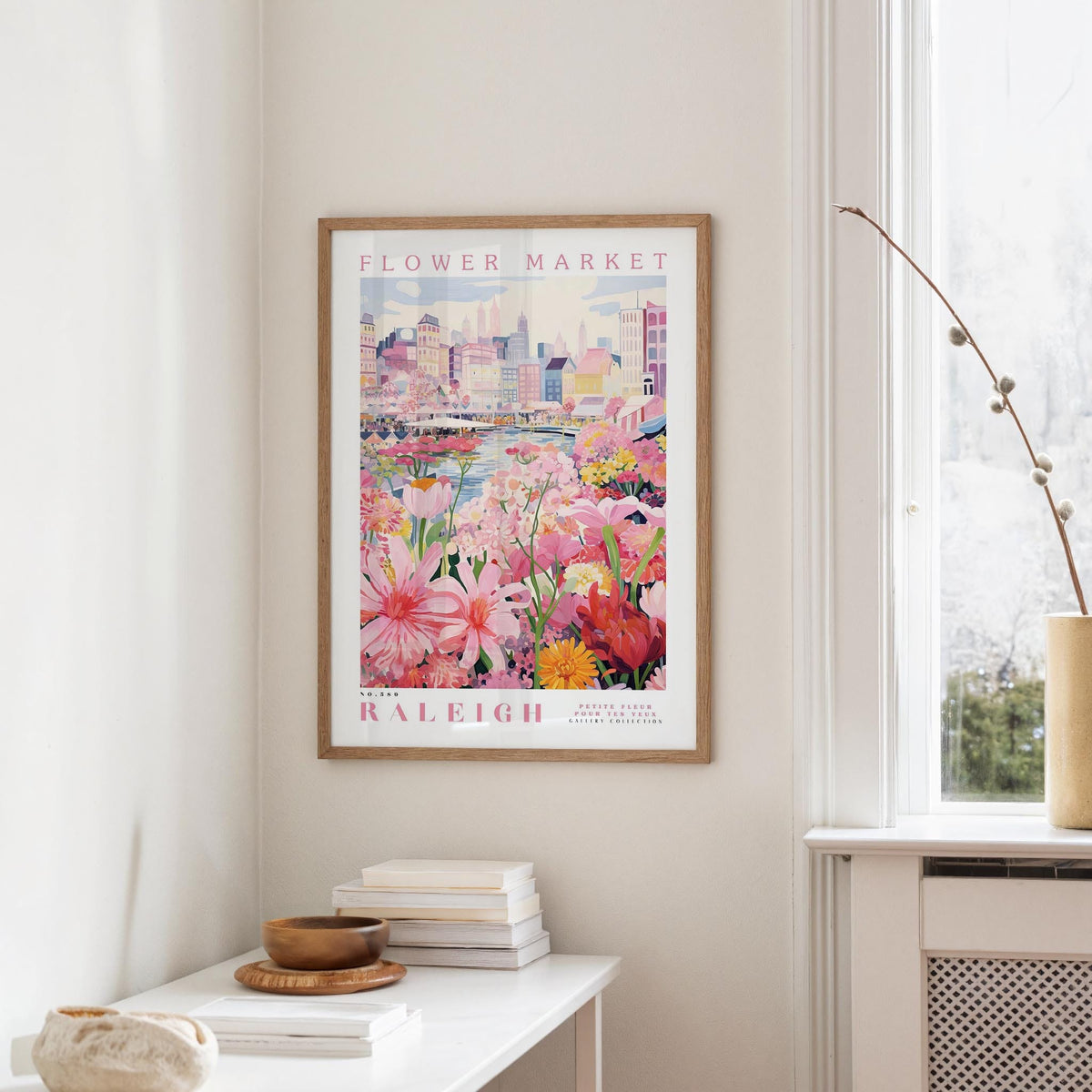 a framed poster of a colorful floral scene hanging on a wall, with a window and a small table with books and a vase in the foreground.