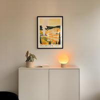 a white cabinet with a plant and a lamp on top, and a framed poster of a scenic landscape hanging on the wall above it.