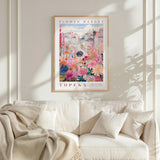 a framed poster of a colorful floral scene hanging on a wall above a white couch with a blanket draped over it.