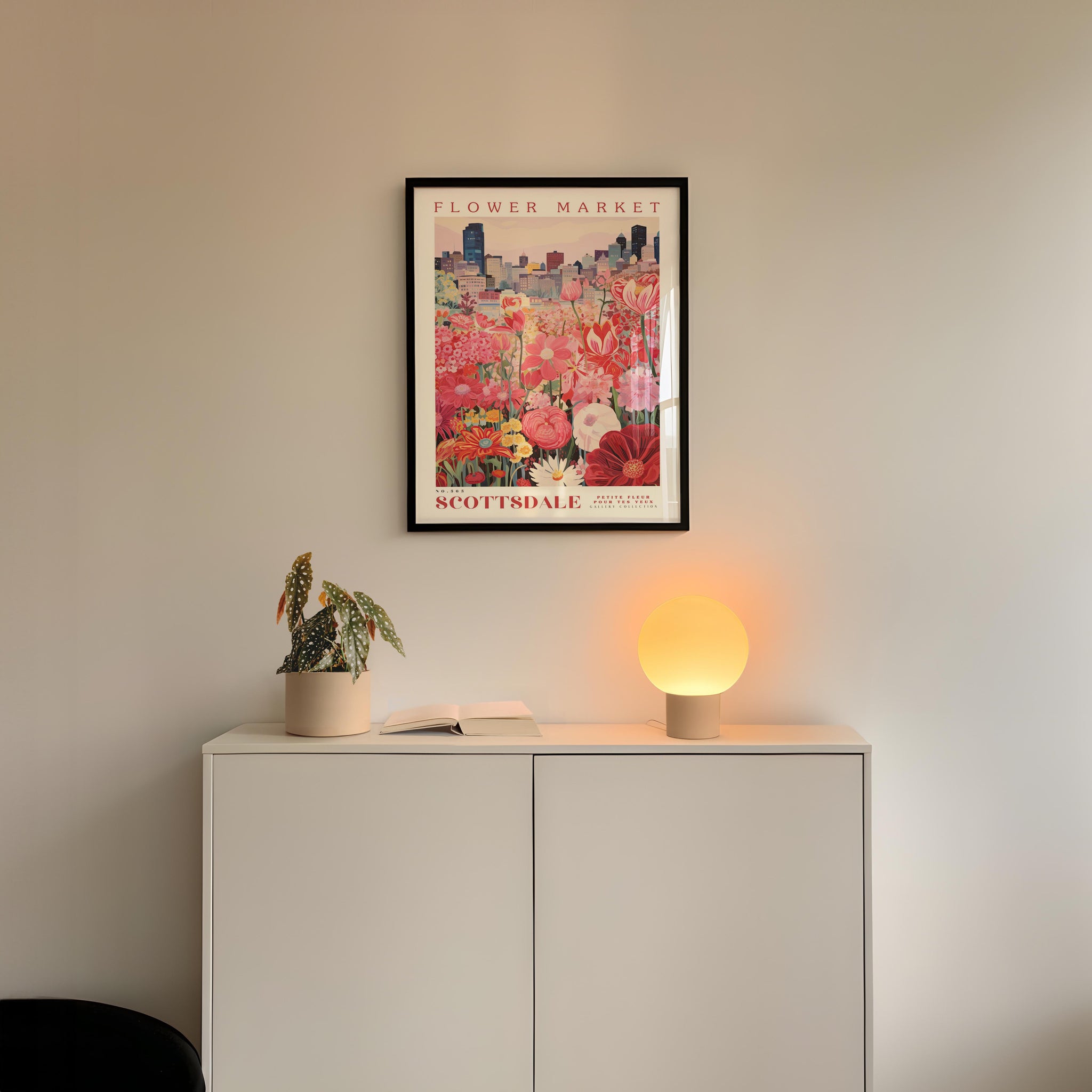 a white cabinet with a potted plant and a lamp on top, set against a wall with a framed poster of a flower market above it.