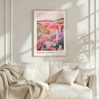 a framed poster of a colorful landscape with flowers hanging on a wall above a white couch with pillows.