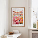 a framed poster of a colorful flower market scene hanging on a wall, with a white table and a vase of flowers in the foreground.