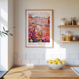 a framed poster of a colorful flower market in Santa Ana, California, hanging on a wall above a wooden counter with a bowl of lemons.