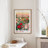 a framed poster of a cityscape with a field of flowers in the foreground, hanging on a wall in a room with a window and a small table with books and a vase.