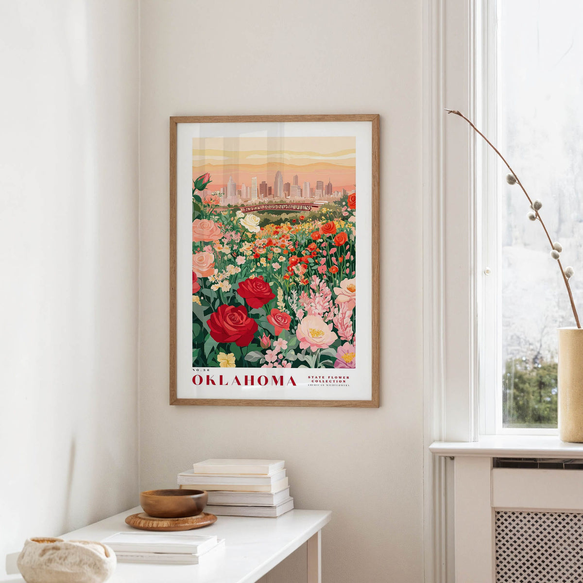 a framed poster of a cityscape with a field of flowers in the foreground, hanging on a wall in a room with a window and a small table with books and a vase.