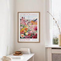 a framed poster of a colorful garden scene hanging on a wall, with a white table and a vase of flowers in the foreground.