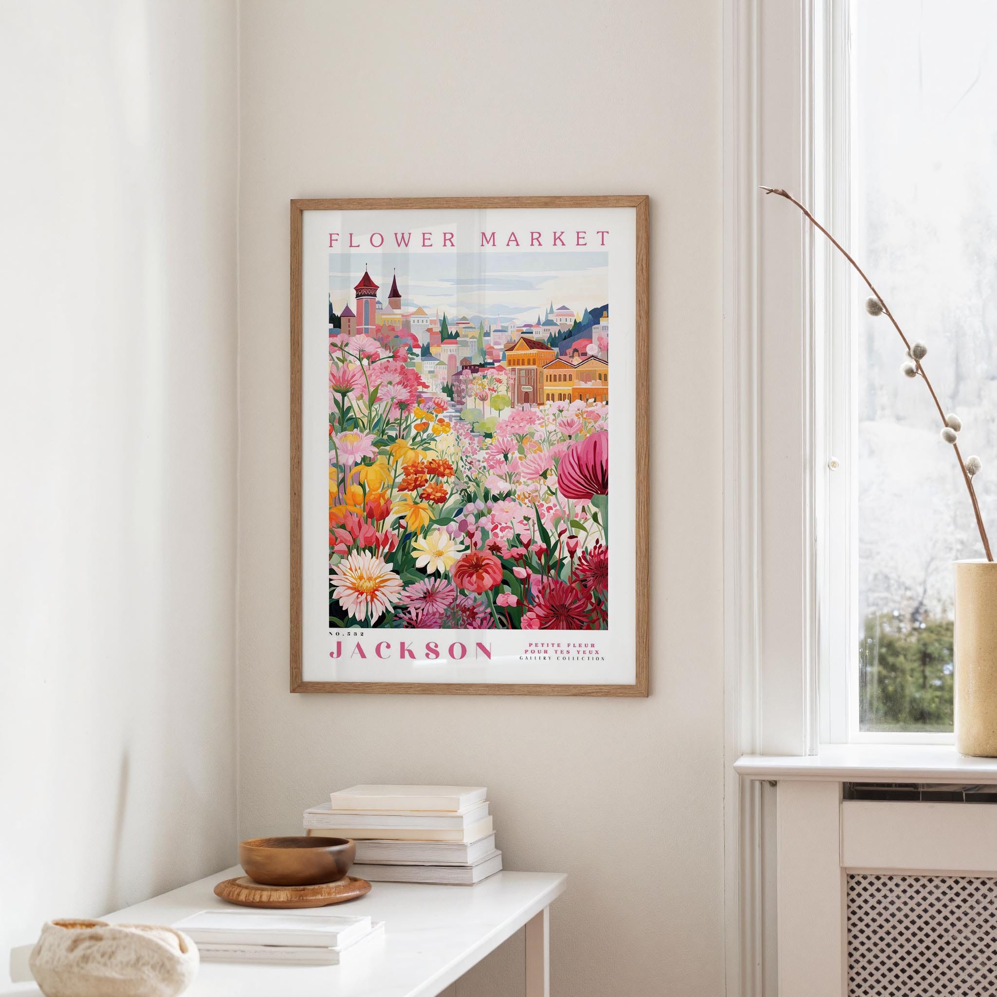 a framed poster of a colorful garden scene hanging on a wall, with a white table and a vase of flowers in the foreground.