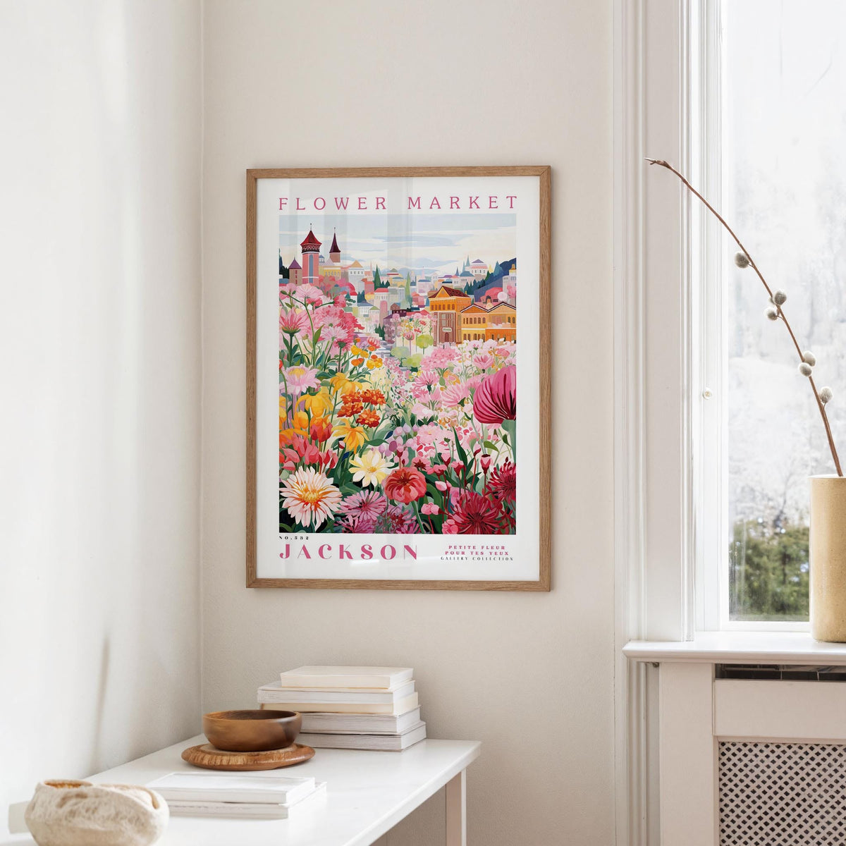 a framed poster of a colorful garden scene hanging on a wall, with a white table and a vase of flowers in the foreground.