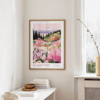 a framed poster of a mountain landscape with a field of pink flowers, hanging on a wall in a room with a white table and a window.