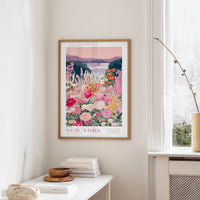 a framed poster of a colorful floral scene hanging on a wall, with a white table and a vase of flowers in the foreground.