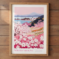 A framed poster depicting a scenic landscape with a mountain range, a small town, and a field of blooming flowers in the foreground.