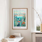 a framed poster of a colorful floral scene hanging on a wall, with a window and a small table with books and a vase in the foreground.