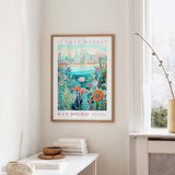 a framed poster of a colorful floral scene hanging on a wall, with a window and a small table with books and a vase in the foreground.