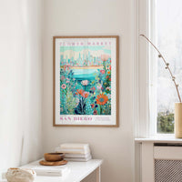 a framed poster of a colorful floral scene hanging on a wall, with a window and a small table with books and a vase in the foreground.