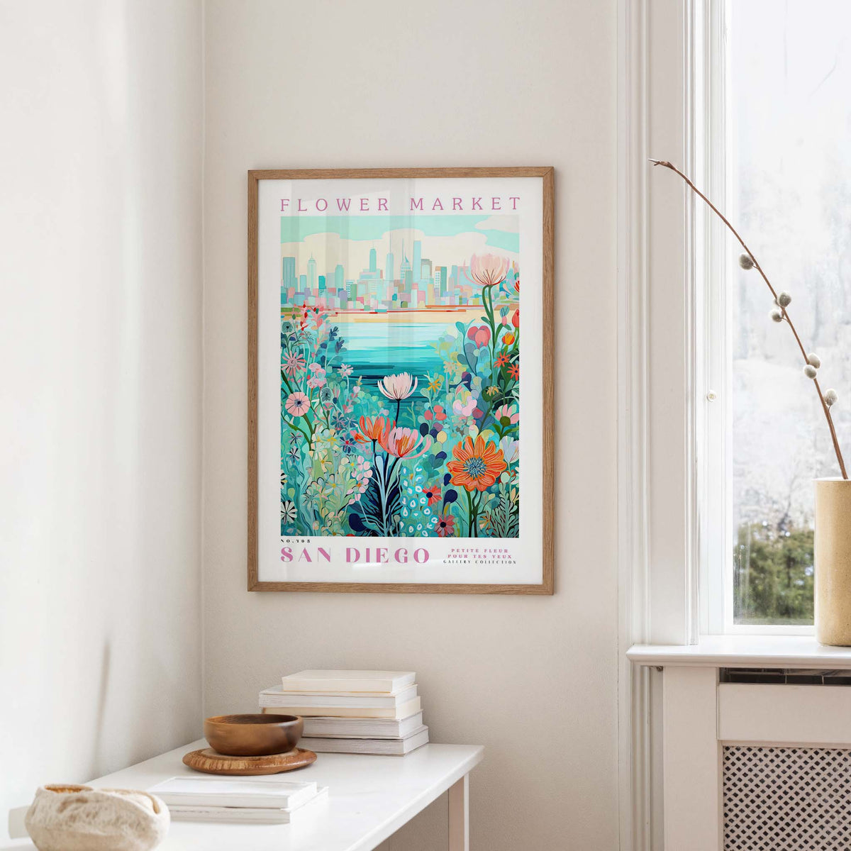 a framed poster of a colorful floral scene hanging on a wall, with a window and a small table with books and a vase in the foreground.