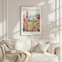 a framed poster of the Golden Gate Bridge and a colorful floral scene, hanging on a wall above a white couch with a blanket draped over it.